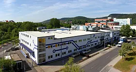 Produktionsgebäude der Jenaer Leiterplatten GmbH in Jena mit weißer Fassade, blauen Leiterbahn-Motiven und mehreren Fenstern. Vor dem zweigeschossigen Gebäude verläuft eine Straße mit parkenden Autos.