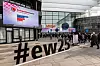 Eingang einer Messehalle mit embedded-world-Logos, Hashtag #ew25 und eintreffenden Besuchern.