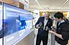 Zwei Männer begutachten Bauteile für gedruckte Elektronik an einem Messestand mit großem Display.