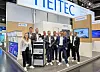 Messestand von Heitec auf einer Fachmesse mit mehreren Personen vor einem Elektroniksystem.