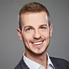 Smiling young professional in a suit jacket against a grey studio background. This is Felix Martin, Research Engineer and AI expert at Tasking.