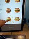 Finger tippt an einem TouchNetix-Bestellterminal in einem Schnellrestaurant auf einen Chicken-Burger.