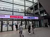 Menschen gehen durch ein verglastes Messefoyer unter einem LED-Banner mit Veranstaltungshinweis.