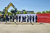 Menschen mit Spaten beim Spatenstich vor Rohde-&-Schwarz-Banner und Bagger