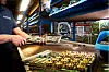 Person zerlegt Leiterplatten an einer Werkbank mit Regalen voller Elektronikbauteile.