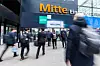 Menschen gehen vor dem Eingang einer Messehalle mit „Mitte“-Schriftzug und „welcome“-Banner.