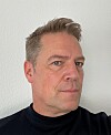 Porträt eines Mannes vor einer hellen Wand, leicht seitlich zur Kamera blickend.