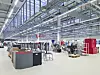 Helle Industriehalle mit Regalen, Maschinen und Paletten unter einer hohen Stahlkonstruktion.