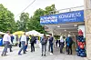 Außenansicht des Veranstaltungsortes beim 30. AUTOMOBIL-ELEKTRONIK KONGRESS 2026 in Ludwigsburg mit großem blauem Welcome-Banner über dem Eingang. Auf dem Banner steht „AUTOMOBIL ELEKTRONIK KONGRESS Welcome!“. Vor dem Gebäude stehen und gehen mehrere Kongressteilnehmer in Businesskleidung, einige mit Rucksäcken oder Lanyards. Links im Bild sind weiße Sonnenschirme und ein kleiner Catering- oder Aktionsstand zu sehen, rechts neben dem Eingang eine Ballondekoration in Rot und Blau. Im Hintergrund befinden sich grüne Bäume und die helle Fassade des Gebäudes. Das Bild zeigt die Ankunftssituation und den Eingangsbereich des Fachkongresses in Ludwigsburg und eignet sich als Schmuckbild für Berichte über den 30. AUTOMOBIL-ELEKTRONIK KONGRESS, Registrierung, Networking, Branchentreffen, Automotive, Elektronik, Konferenzstandort Ludwigsburg, Veranstaltungseingang und Kongressatmosphäre.