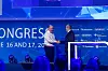 Ricky Hudi und Alfred Vollmer stehen auf der Bühne des 30. AUTOMOBIL-ELEKTRONIK KONGRESSES 2026 in Ludwigsburg und geben sich bei der symbolischen Übergabe des Chairman-Postens im Advisory Board die Hand. Links im Bild steht Ricky Hudi im weißen Hemd, rechts Alfred Vollmer im dunklen Anzug. Beide befinden sich vor dem blauen Bühnenhintergrund des Kongresses mit großem Schriftzug „Kongress“, dem Veranstaltungsdatum 16. und 17. Juni 2026 sowie Sponsorlogos von Microsoft, Qualcomm, Synopsys, Texas Instruments und T-Systems. Im Vordergrund sind die Köpfe des Publikums zu sehen, die Bühne ist in blau-violettes Licht getaucht. Das Bild dokumentiert den Wechsel an der Spitze des Advisory Boards beim AEK, die Verabschiedung von Ricky Hudi als Chairman und die Übergabe an Alfred Vollmer. Geeignet für Berichte über den 30. AUTOMOBIL-ELEKTRONIK KONGRESS, AEK 2026, Ricky Hudi, Alfred Vollmer, Advisory Board, Staffelstabübergabe, Kongresseröffnung, Automotive-Konferenz, Branchentreffen, Elektronikindustrie und Zukunft der Fahrzeugtechnologien.