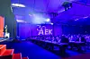 Blick in den Konferenzsaal des AEK 2026 in Ludwigsburg während eines Vortrags. Links steht ein Speaker auf der Bühne, rechts sitzen die Teilnehmer an langen Tischen und verfolgen das Programm. Im Hintergrund ist groß das beleuchtete Jubiläumslogo „30 AEK“ auf dem Vorhang zu sehen. Die Bühne und der Saal sind in blau-violettes Licht getaucht, auf den Tischen stehen Wasserflaschen, Gläser und Arbeitsmaterialien. Das Bild zeigt die Atmosphäre des 30. AUTOMOBIL-ELEKTRONIK KONGRESSES, das Jubiläum des AEK, das Fachpublikum im Saal und die Inszenierung der Veranstaltung rund um Automotive, Elektronik, Software, KI und Fahrzeugtechnologien.