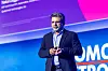 Nakul Duggal von Qualcomm Technologies steht auf der Bühne des 30. AUTOMOBIL-ELEKTRONIK Kongresses 2026 in Ludwigsburg und hält einen Vortrag über KI im Fahrzeug, Software-defined Vehicles und neue digitale Kundenerlebnisse. Er trägt einen dunklen Anzug und ein blaues Hemd, spricht mit Headset-Mikrofon und hält einen Presenter in der Hand. Im Hintergrund ist eine violett-pinke Präsentationsfolie zu sehen, auf der unscharf der Name Nakul Duggal sowie seine Rolle als Executive Vice President und Group General Manager Automotive, Industrial and Embedded IoT bei Qualcomm Technologies zu erkennen sind. Die Bühne ist blau und violett ausgeleuchtet, im Hintergrund sind Teile des Kongressbrandings sichtbar. Das Bild eignet sich für Berichte über Qualcomm, Nakul Duggal, KI im Auto, Fahrzeug-SoCs, Software-defined Vehicles, Automotive Computing, Edge AI, digitale Fahrzeugplattformen, Kundenerlebnis im Auto und den 30. AUTOMOBIL-ELEKTRONIK Kongress in Ludwigsburg.