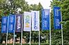 Flaggen vor dem Veranstaltungsort des 30. AUTOMOBIL-ELEKTRONIK Kongresses 2026 in Ludwigsburg. Mehrere blaue Kongressflaggen sowie Partnerflaggen von Ansys und Qualcomm stehen vor grünen Bäumen und markieren den Außenbereich der Veranstaltung. Die Aufnahme zeigt die Präsenz des Kongresses bereits vor dem Gebäude und eignet sich als Schmuckbild für Berichte über den AEK, den AUTOMOBIL-ELEKTRONIK Kongress, Partner und Sponsoren, Automotive-Community, Branchentreff, Kongressstandort Ludwigsburg, Automotive Computing, Elektronik, Software-defined Vehicles, KI im Auto und Fahrzeugtechnologien.