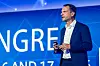 Walter Glück von Leoni steht auf der Bühne des 30. AUTOMOBIL-ELEKTRONIK Kongresses 2026 in Ludwigsburg und hält einen Vortrag über künftige E/E-Architekturen, Kabelbaumkomplexität, automatisierte Fertigung und neue Lieferkettenmodelle. Er trägt einen dunklen Anzug und ein weißes Hemd, spricht mit Headset-Mikrofon und hält einen Presenter in der Hand. Im Hintergrund ist der blaue Bühnenhintergrund des Kongresses mit großen weißen Buchstaben und grafischen Netzwerkelementen zu sehen. Das Bild eignet sich für Beiträge über Walter Glück, Leoni, Wiring Harness, Kabelbaum, E/E-Architektur, Zonenarchitektur, automatisierte Kabelbaumfertigung, Smart Manufacturing, modulare Fahrzeugarchitekturen, Lieferkette, Produktion, Standardisierung, Software-defined Vehicles, Automotive Engineering und den 30. AUTOMOBIL-ELEKTRONIK Kongress in Ludwigsburg.