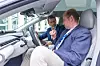 Zwei Teilnehmer des 30. AUTOMOBIL-ELEKTRONIK Kongresses 2026 sitzen im Innenraum eines ausgestellten Fahrzeugs und sprechen über das Cockpit, die Displays und die Fahrzeugtechnik. Links sitzt ein Mann im blauen Sakko auf dem Fahrersitz und deutet auf den Innenraum, rechts sitzt ein weiterer Teilnehmer im hellen Sitzbereich des Fahrzeugs. Zu sehen sind Lenkrad, Armaturentafel, Infotainment-Displays, helles Interieur und große Fensterflächen des Autos. Die Szene zeigt, dass die ausgestellten Fahrzeuge beim AEK nicht nur Exponate im Außenbereich sind, sondern auch zum Einsteigen, Ausprobieren und Diskutieren einladen. Das Bild eignet sich für Beiträge über den AUTOMOBIL-ELEKTRONIK Kongress, Fahrzeugausstellung, Automotive UX, Innenraumtechnik, Cockpit, Infotainment, Software-defined Vehicles, digitale Fahrzeugfunktionen, E/E-Architekturen, Kundenerlebnis im Auto, BMW-Fahrzeuge, Elektromobilität und den fachlichen Austausch in Ludwigsburg.