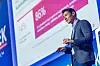 Dominik Wee, Corporate Vice President Forward-Deployed Engineering bei Microsoft, steht auf der Bühne des 30. AUTOMOBIL-ELEKTRONIK Kongresses 2026 in Ludwigsburg und hält einen Vortrag über KI in der Softwareentwicklung, spec-driven development, Agenten, Automotive Software und die Zukunft des Programmierens. Er trägt ein graues Sakko, ein hellblaues Hemd, Jeans und ein Headset-Mikrofon, hält einen Presenter in der Hand und spricht vor einer großen Präsentationsfolie mit Microsoft-Branding. Auf der Folie sind Kennzahlen zur Nutzung von KI in Entwicklungsprozessen zu sehen, darunter eine hervorgehobene Prozentzahl. Die Bühne ist blau ausgeleuchtet, links ist das AEK-Branding teilweise sichtbar. Das Bild eignet sich für Beiträge über Dominik Wee, Microsoft, KI-Agenten, generative KI, Softwareentwicklung im Auto, Automotive Software Engineering, V-Modell, Spezifikationen, Compliance, Domain Expertise, Software-defined Vehicles und den AUTOMOBIL-ELEKTRONIK Kongress in Ludwigsburg.