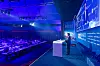Michael Muth, Executive Advisor von Audi China, steht auf der Bühne des 30. AUTOMOBIL-ELEKTRONIK Kongresses 2026 in Ludwigsburg und eröffnet den zweiten Konferenztag mit einem Vortrag über die Elektrifizierung, Digitalisierung und Innovationsdynamik des chinesischen Automobilmarkts. Das Foto zeigt eine weite Perspektive aus dem Veranstaltungssaal: Links sitzt das Publikum an langen Konferenztischen, rechts steht Michael Muth an einem weißen Rednerpult vor der großflächig blau ausgeleuchteten Bühne. Auf der Bühnenwand sind das Branding des AUTOMOBIL-ELEKTRONIK Kongresses, das Veranstaltungsdatum 16. und 17. Juni 2026 sowie zahlreiche Sponsorenlogos zu sehen, darunter Qualcomm, Synopsys, Microsoft und T-Systems. Auf der großen Präsentationsfläche ist Michael Muth zusätzlich als Live-Kamerabild eingeblendet; daneben sind Folieninhalte zur Entwicklung in China und zur Rolle lokaler Technologie- und Innovationsökosysteme erkennbar. Die Bühne ist in kräftigen Blau- und Violetttönen beleuchtet, im Vordergrund und im Saal ist die konzentrierte Atmosphäre des zweiten Kongresstages zu sehen. Das Bild eignet sich für Beiträge über Michael Muth, Audi China, Chinas Automobilmarkt, Elektrifizierung in China, digitale Fahrzeugökosysteme, lokale Entwicklungsprozesse, Software-defined Vehicles, AI-defined Vehicles, Physical AI, Automotive Innovation, chinesische Tech-Ökosysteme, globale Fahrzeugentwicklung, OEM-Strategien, den AUTOMOBIL-ELEKTRONIK Kongress 2026 und den AEK in Ludwigsburg.