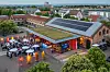 Luftaufnahme des Abendevents beim 30. AUTOMOBIL-ELEKTRONIK Kongress 2026 in Ludwigsburg. Zu sehen ist ein historisches Veranstaltungsgebäude mit Backsteinfassade, Glasfront, begrüntem Dach und Solarpaneelen auf dem Dach. Vor dem Gebäude stehen zahlreiche Teilnehmer in Gruppen zusammen, unter weißen Sonnenschirmen und neben dekorativen Leuchtelementen. An der Glasfront sind AEK-Branding und Veranstaltungsdisplays zu erkennen, während die Umgebung mit Bäumen, Gebäuden und dem Blick über Ludwigsburg den Rahmen bildet. Das Bild vermittelt die Größe und Atmosphäre des Networking-Abends und eignet sich für Beiträge über das AEK-Abendevent, Networking, Kongresscommunity, Veranstaltungsort Ludwigsburg, Abendempfang, Teilnehmer, Speaker, Partner und den AUTOMOBIL-ELEKTRONIK Kongress 2026.