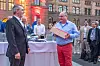 Ricky Hudi wird beim Abendevent des 30. AUTOMOBIL-ELEKTRONIK Kongresses 2026 in Ludwigsburg im Kreis der AEK-Community verabschiedet. Im Vordergrund steht Ricky Hudi im hellblauen Hemd und roter Hose und hält ein Geschenkpaket in den Händen. Ihm gegenüber steht ein Mann im dunklen Anzug mit Mikrofon, während mehrere Teilnehmer im Hintergrund lächeln und den Moment verfolgen. Auf dem Tisch sind Tellerstapel, ein Mikrofon, ein Modellauto und eine 30-Jahre-AEK-Dekoration zu sehen. Die Szene findet im Außenbereich des Abendempfangs vor einer historischen Backsteinfassade statt und ist in warmes Abendlicht getaucht. Das Bild eignet sich für Beiträge über Ricky Hudi, seine Verabschiedung, 30 Jahre AEK, den Wechsel im Advisory Board, die AEK-Community, das Abendevent, persönliche Würdigung, Dank und den AUTOMOBIL-ELEKTRONIK Kongress in Ludwigsburg