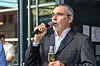 Ein Mann im dunklen Anzug hält beim Abendevent des 30. AUTOMOBIL-ELEKTRONIK Kongresses 2026 in Ludwigsburg ein Mikrofon in der rechten Hand und ein Sektglas in der linken Hand. Er steht vor einer Glasfront und spricht zu den Teilnehmern des Networking-Events. Die Szene wirkt feierlich und zugleich ungezwungen, passend zum Ausklang des ersten Kongresstages. Das Bild zeigt die Atmosphäre des Abendprogramms, bei dem Teilnehmer, Speaker, Partner und Veranstalter zusammenkommen, um Gespräche fortzusetzen, auf den Kongress anzustoßen und besondere Momente des AEK zu würdigen. Es eignet sich für Beiträge über das AEK-Abendevent, Networking, Kongressatmosphäre, Abendempfang, Community, persönliche Begegnungen und den AUTOMOBIL-ELEKTRONIK Kongress in Ludwigsburg.