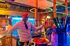 Ein Barkeeper steht beim Abendevent des 30. AUTOMOBIL-ELEKTRONIK Kongresses 2026 in Ludwigsburg hinter einer Bar und hält eine Flasche in der Hand. Die Szene ist in farbiges Licht in Rot, Blau und Grün getaucht, im Hintergrund sind Glasflächen, Leuchtelemente und Barzubehör zu sehen. Im Vordergrund stehen unscharf Becher, Flaschen, Strohhalme und Zutaten bereit. Das Bild zeigt die entspannte Atmosphäre des Abendempfangs nach einem intensiven Kongresstag und eignet sich für Beiträge über das AEK-Abendevent, Networking, Bar, Getränke, lockere Gespräche, Kongressausklang, Teilnehmererlebnis, Community und den AUTOMOBIL-ELEKTRONIK Kongress in Ludwigsburg.