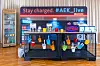Die AEK Fueling Station beim 30. AUTOMOBIL-ELEKTRONIK Kongress 2026 in Ludwigsburg ist als dekorativer Pausen- und Versorgungsbereich aufgebaut. Über der Theke leuchtet der Schriftzug „Stay charged. #AEK_live“, daneben ist das Branding von Automotive Digital Transformation zu sehen. Auf und hinter der Theke stehen Getränke, Snacks, Süßigkeiten, Becher und Zeitschriften, links befindet sich ein Kühlschrank mit Softdrinks und Getränkedosen. Bunte kleine Eimer hängen an einem schwarzen Gitter vor der Theke, rechts leuchtet ein „Open“-Schild. Das Bild eignet sich für Beiträge über Pausenversorgung, Teilnehmerkomfort, AEK Fueling Station, Snacks, Getränke, Kongresspausen, Live-Berichterstattung, #AEK_live, Automotive Digital Transformation und den AUTOMOBIL-ELEKTRONIK Kongress in Ludwigsburg.