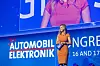 Maxi Sarwas steht als Moderatorin auf der Bühne des 30. AUTOMOBIL-ELEKTRONIK Kongresses 2026 in Ludwigsburg und führt durch das Konferenzprogramm. Sie trägt ein orangefarbenes Kleid, ein Headset-Mikrofon und hält Moderationskarten mit AEK-Branding in den Händen. Hinter ihr ist die großflächige blaue Bühnenwand mit dem Schriftzug AUTOMOBIL-ELEKTRONIK Kongress, dem 30-Jahre-Jubiläumslogo und dem Veranstaltungsdatum 16. und 17. Juni 2026 zu sehen. Oberhalb der Bühne erscheint Maxi Sarwas zusätzlich als Live-Kamerabild auf der großen Leinwand. Die Szene zeigt eine professionelle Kongressmoderation in blauem Bühnenlicht und eignet sich für Beiträge über Maxi Sarwas, Moderation, Konferenzprogramm, Live-Bühne, Automotive-Event, AEK 2026, 30 Jahre AUTOMOBIL-ELEKTRONIK Kongress, Fachkongress in Ludwigsburg, Fahrzeugelektronik, Software-defined Vehicles, KI im Auto und Automotive Networking.