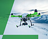 Grüne Drohne mit Kamera schwebt über einer verschneiten Gebirgslandschaft.