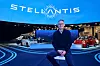 Ned Curic, EVP, Chief Engineering Officer and CTO bei Stellantis, sitzt auf einem Hocker vor dem blau beleuchteten Stellantis-Messestand mit großem Stellantis-Logo und mehreren ausgestellten Fahrzeugen. Das Bild zeigt Curic in einem dunklen Pullover über einem Hemd vor einer modernen Automotive-Inszenierung mit Fahrzeugen, Lichtlinien und digitalen Displays. Das Motiv passt zu einem Interview über Stellantis, Software-defined Vehicle, STLA Brain, STLA SmartCockpit, digitale Fahrzeugarchitekturen, Customer Experience, Cockpit-Bedienung, Personalisierung, globale Softwareplattformen, regionale Anforderungen, Markenidentität, vernetzte Dienste, Fahrzeugsoftware, Automotive Electronics und die Frage, wie digitale Funktionen im Auto einfacher und kundenorientierter gestaltet werden können.