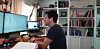 Mann sitzt am Schreibtisch vor zwei Monitoren in einem Homeoffice mit Bücherregalen im Hintergrund.