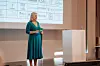 Eine Frau steht auf einer Bühne vor einer Projektionswand mit Logos und hält Karten in der Hand.