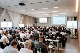 Großer Konferenzsaal mit vielen sitzenden Teilnehmenden, Leinwänden und einer Bühne im Hintergrund.