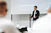 Ein Mann hält einen Vortrag vor einer Projektionsleinwand in einem Konferenzraum; Zuhörer sind unscharf im Vordergrund zu sehen.
