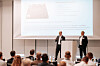 Zwei Männer stehen vor einer Projektionsleinwand und sprechen vor sitzendem Publikum bei einer Fachkonferenz.