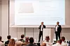 Zwei Männer stehen vor einer Projektionsleinwand und sprechen vor sitzendem Publikum bei einer Fachkonferenz.