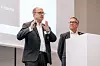 Zwei Männer in Anzügen stehen vor einer Leinwand und einem Rednerpult bei einer Fachveranstaltung.