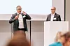 Zwei Männer stehen auf einer Bühne vor einem Publikum in einem Konferenzraum.