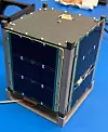 Fertig montierter BOHR-CubeSat von City Labs auf einer blauen Arbeitsfläche während der Vorbereitung auf seinen Weltraumeinsatz. Der kompakte Kleinsatellit besitzt ein würfelförmiges graues Metallgehäuse mit verstärkten Ecken, zahlreichen Schraubverbindungen und dunklen Solarzellen auf den sichtbaren Seitenflächen. Auf der Oberseite befinden sich eine geschlossene Metallplatte, Befestigungselemente und ein kleiner elektrischer Anschluss. Die Solarflächen liefern die Energie für den Betrieb des Satellitenbusses. Im Inneren des CubeSats ist eine separate experimentelle Nutzlast mit der betavoltaischen NanoTritium-Batterie untergebracht. Diese erzeugt aus dem radioaktiven Zerfall des Wasserstoffisotops Tritium über eine Halbleiterstruktur kontinuierlich geringe Mengen elektrischen Stroms. Die BOHR-Mission soll zeigen, wie zuverlässig die langlebige Nuklearbatterie den Start und den Betrieb unter Vakuum, Strahlung und starken Temperaturschwankungen im Erdorbit übersteht.