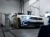 Weißer Volkswagen auf einem Rollenprüfstand in einer Testhalle mit eingeschaltetem Scheinwerfer.