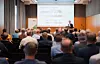 Rückansicht eines gut besuchten Vortragssaals während der 33. FED-Konferenz 2025 in Lübeck. Zahlreiche Teilnehmer sitzen in mehreren Reihen und verfolgen einen Fachvortrag. Vorne steht ein Referent an einem Rednerpult neben einer großen Projektionsleinwand, auf der eine Präsentation zum Thema „Problematik im Designflow“ mit Grafiken und Prozessdarstellungen aus dem Elektronikdesign gezeigt wird. Links neben der Leinwand befindet sich ein Roll-up des Fachverbands Elektronikdesign und -fertigung (FED). Der modern ausgestattete Konferenzraum verfügt über Holzverkleidungen, hohe Fenster mit Vorhängen und eine professionelle Veranstaltungstechnik. Das Bild dokumentiert die Vortragsatmosphäre und den fachlichen Austausch auf der FED-Konferenz 2025 in Lübeck.