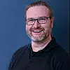 Lächelnder Mann mit Brille und Bart vor blauem Hintergrund. Markus Rayen von Drews electronic