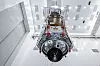 Untersicht des nahezu vollständig montierten Nancy Grace Roman Space Telescope in einer hohen, hell beleuchteten Reinraumhalle des Kennedy Space Center in Florida. Das große Weltraumobservatorium hängt senkrecht an mehreren dünnen Tragseilen und wird mit einem Hallenkran zu einem speziellen Arbeitsstand gehoben. Die Aufnahme entstand am 26. Juni 2026 im Payload Hazardous Servicing Facility während der Vorbereitung des NASA-Weltraumteleskops auf seinen Start.

Roman füllt den mittleren Teil des querformatigen Fotos fast vollständig aus und wird aus einer tiefen Perspektive von schräg unten gezeigt. Dadurch wirkt das Observatorium langgestreckt und technisch komplex. Der untere, dem Betrachter zugewandte Bereich besitzt einen großen kreisförmigen Anschlussring. Innerhalb dieses Rings sind zahlreiche Leitungen, Kabel, Rohre, Steckverbindungen, elektronische Baugruppen und metallische Strukturelemente sichtbar. Vor der runden Öffnung befindet sich ein großes schwarzes, kreisförmiges Bauteil mit einer in Segmente unterteilten Oberfläche. Dünne weiße Seile oder Leitungen verlaufen davor und verbinden die Baugruppe mit den Hebevorrichtungen.

Der zentrale Körper des Roman Space Telescope ist überwiegend mit silbern reflektierender mehrlagiger Isolationsfolie verkleidet. Die Folie ist an vielen Stellen gefaltet und bildet eine unregelmäßige, metallisch glänzende Oberfläche. Sie umschließt große Teile des Raumfahrzeugs und dient sichtbar als thermische Verkleidung der darunterliegenden Technik. Auf und neben der Isolierung sitzen mehrere rote rechteckige Schutz-, Halte- oder Befestigungselemente. Weitere rote Bauteile sind entlang des unteren kreisförmigen Rings und an den seitlichen Strukturen angebracht.

Oberhalb des silbern verkleideten Raumfahrzeugkörpers erstreckt sich eine längliche, dunkelbraune bis schwarze Fläche aus mehreren rechteckigen Abschnitten. Auf dieser Struktur sind helle Befestigungspunkte, kleine technische Komponenten und orangefarbene Leitungen oder Markierungen zu erkennen. Die Fläche verjüngt sich perspektivisch zum oberen Bildrand. Seitlich verlaufen große metallische Streben und schräg angeordnete Abdeckungen. Am oberen Ende ist eine breite Hebestruktur mit weiteren Tragseilen befestigt. Transparente Schutzfolien hängen links und rechts hinter dem oberen Bereich des Teleskops und trennen die technische Arbeitszone teilweise von der Umgebung.

Die Reinraumhalle besitzt weiße Wände und eine sehr hohe weiße Decke. Zahlreiche rechteckige Leuchten, Lüftungsfelder und graue Deckenpaneele sind regelmäßig über den Raum verteilt. Links befindet sich ein sehr großes geschlossenes Rolltor mit horizontalen Metalllamellen. Rechts oben verläuft ein weißer Hallenkran; auf einem seiner Bauteile ist die Aufschrift „Ederer“ sichtbar. Mehrere dünne Kranseile reichen von der Deckenkonstruktion bis zum Weltraumteleskop. Personen sind in diesem Bildausschnitt nicht zu sehen.

Die Aufnahme vermittelt die Größe und den Integrationsaufwand des Nancy Grace Roman Space Telescope kurz vor dem Start. Hinter den sichtbaren Strukturen und Isolationsschichten arbeitet ein technisches Gesamtsystem aus 18 gekühlten H4RG-10-Infrarotsensoren mit rund 302 Millionen Pixeln, Bordelektronik, Flugsoftware, Reaktionsrädern, thermischer Regelung, aktiver Wellenfrontkorrektur und einer Datenarchitektur für bis zu 11 Tbit komprimierte Beobachtungsdaten pro Tag. Das Bild zeigt das NASA-Weltraumobservatorium während einer der letzten bodenseitigen Arbeitsphasen, bevor es große Himmelsfelder im sichtbaren und nahen Infrarotbereich untersuchen soll.