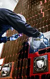 Hochformatige Nahaufnahme eines Technikers bei der Reinigung und Kontrolle eines Solarpaneels des Nancy Grace Roman Space Telescope im Payload Hazardous Servicing Facility des Kennedy Space Center in Florida. Die Person steht links und vor dem großflächigen Solar Array Sun Shield. Sie trägt dunkelblaue Reinraumkleidung, eine weiße Haube, einen Gesichtsschutz sowie hellblaue Schutzhandschuhe. Der Kopf und große Teile des Oberkörpers sind nach vorn geneigt, sodass das Gesicht fast vollständig verdeckt ist.

Eine behandschuhte Hand hält eine kleine Lampe oder Taschenleuchte, die violettweißes Licht auf die Oberfläche des Paneels richtet. Die andere Hand arbeitet am rechten Rand eines silbrig reflektierenden Feldes. Auf der glänzenden Oberfläche spiegeln sich die Hand, die Lichtquelle und Teile der Reinraumumgebung. Die Arbeitsposition und die gerichtete Beleuchtung deuten auf eine sorgfältige Sichtprüfung und Reinigung hin.

Der größte Teil des Hintergrunds besteht aus vielen rechteckigen, dunkelbraunen bis nahezu schwarzen Solarzellen. Sie sind in einem regelmäßigen Raster auf einer rot-orangefarbenen Trägerstruktur angeordnet. Schmale rote Zwischenräume und Befestigungsbereiche trennen die einzelnen Zellen voneinander. Einige Bereiche wirken aufgrund der Spiegelung heller, während andere fast schwarz erscheinen.

Im unteren Teil des Paneels sind mehrere technische Anbauteile sichtbar. Dazu gehören zwei helle, annähernd kegelförmige Komponenten in quadratischen Metallfassungen, gelbe Leitungen, Schrauben sowie rote und schwarze Strukturflächen. Rechts neben dem Techniker befindet sich ein rechteckiger Bereich mit silbrig glänzender Oberfläche und feinem Rastermuster.

Das Foto zeigt die empfindliche Oberfläche des Solar Array Sun Shield kurz vor dem Start des Roman Space Telescope. Die Aufnahme verdeutlicht den Aufwand der Kontaminationskontrolle bei Raumfahrthardware. Staub, Partikel oder Rückstände müssen entfernt werden, bevor die Paneele im All Sonnenlicht in elektrische Energie umwandeln und zugleich das Observatorium gegen direkte Sonneneinstrahlung abschirmen.