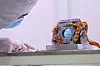 Querformatige Nahaufnahme eines Fast Steering Mirror für das Coronagraph Instrument des Nancy Grace Roman Space Telescope in einer hellen Reinraumumgebung. Rechts in der Bildmitte steht die kompakte optomechanische Baugruppe auf einer massiven silberfarbenen Metallhalterung. Im Zentrum der Baugruppe befindet sich eine runde spiegelnde Fläche mit bläulicher Reflexion. Der Spiegel ist in eine dunkelgraue quadratische Fassung mit abgeschrägten Ecken eingebaut. Rund um die Fassung sind Schrauben, kleine Halterungen, Gelenke und präzise bearbeitete Metallteile sichtbar.

Mehrere Bündel orange- bis kupferfarbener isolierter Leitungen verlaufen von beiden Seiten über und um die Spiegelbaugruppe. Die Kabel sind teilweise zu Schleifen gelegt und mit weißen sowie blauen Bändern zusammengebunden. Sie verbinden die bewegliche optische Einheit mit der elektrischen Ansteuerung und den außerhalb des Bildausschnitts liegenden Systemen. Unterhalb der Fassung sitzen zylindrische Abstandshalter und weitere feinmechanische Bauteile auf einer rechteckigen Grundplatte.

Links ist ein Ingenieur oder Techniker in Reinraumbekleidung zu sehen. Die Person trägt eine weiße Haube, einen hellblauen Mund-Nasen-Schutz und weiße Handschuhe. Nur ein Teil des Gesichts ist sichtbar. Eine behandschuhte Hand liegt nahe der Arbeitsfläche, ohne die Spiegelbaugruppe zu berühren. Der Hintergrund besteht aus einer hellen, weich ausgeleuchteten Reinraumwand.

Das Foto zeigt die geringe Größe und den präzisen mechanischen Aufbau des Fast Steering Mirror. Die sichtbare Verkabelung, die stabile Fassung und der kreisförmige Spiegel verdeutlichen die Verbindung von elektromechanischer Ansteuerung und optischer Feinregelung im Roman Space Telescope. Das Bauteil soll sehr kleine Bewegungen ausgleichen, damit der Koronograf das Licht eines Sterns möglichst stabil unterdrücken kann.