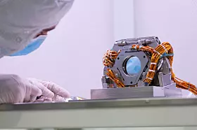 Querformatige Nahaufnahme eines Fast Steering Mirror für das Coronagraph Instrument des Nancy Grace Roman Space Telescope in einer hellen Reinraumumgebung. Rechts in der Bildmitte steht die kompakte optomechanische Baugruppe auf einer massiven silberfarbenen Metallhalterung. Im Zentrum der Baugruppe befindet sich eine runde spiegelnde Fläche mit bläulicher Reflexion. Der Spiegel ist in eine dunkelgraue quadratische Fassung mit abgeschrägten Ecken eingebaut. Rund um die Fassung sind Schrauben, kleine Halterungen, Gelenke und präzise bearbeitete Metallteile sichtbar.

Mehrere Bündel orange- bis kupferfarbener isolierter Leitungen verlaufen von beiden Seiten über und um die Spiegelbaugruppe. Die Kabel sind teilweise zu Schleifen gelegt und mit weißen sowie blauen Bändern zusammengebunden. Sie verbinden die bewegliche optische Einheit mit der elektrischen Ansteuerung und den außerhalb des Bildausschnitts liegenden Systemen. Unterhalb der Fassung sitzen zylindrische Abstandshalter und weitere feinmechanische Bauteile auf einer rechteckigen Grundplatte.

Links ist ein Ingenieur oder Techniker in Reinraumbekleidung zu sehen. Die Person trägt eine weiße Haube, einen hellblauen Mund-Nasen-Schutz und weiße Handschuhe. Nur ein Teil des Gesichts ist sichtbar. Eine behandschuhte Hand liegt nahe der Arbeitsfläche, ohne die Spiegelbaugruppe zu berühren. Der Hintergrund besteht aus einer hellen, weich ausgeleuchteten Reinraumwand.

Das Foto zeigt die geringe Größe und den präzisen mechanischen Aufbau des Fast Steering Mirror. Die sichtbare Verkabelung, die stabile Fassung und der kreisförmige Spiegel verdeutlichen die Verbindung von elektromechanischer Ansteuerung und optischer Feinregelung im Roman Space Telescope. Das Bauteil soll sehr kleine Bewegungen ausgleichen, damit der Koronograf das Licht eines Sterns möglichst stabil unterdrücken kann.