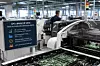 Automatisierte Maschine in einer Fabrikhalle mit sichtbaren Leiterplatten und einem Bediener im Hintergrund.