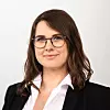 Frau mit Brille und schwarzem Blazer vor hellem Hintergrund.