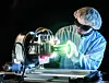 Techniker von Jena-Optronik bei der Prüfung eines optischen Sensorsystems in einem Labor für Raumfahrttechnik. Der Mitarbeiter trägt Reinraumkleidung, Haarnetz, Handschuhe und Schutzbrille und arbeitet an einem metallischen Prüfaufbau mit intensiv grünem Lichtstrahl. Jena-Optronik aus Jena liefert für das Nancy Grace Roman Space Telescope drei ASTRO-APS-Sternsensoren zur präzisen Lage- und Orientierungsbestimmung des Weltraumteleskops.