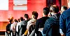 Rückenansicht mehrerer Zuhörer in einem Konferenzsaal mit roter Bühne im Hintergrund.