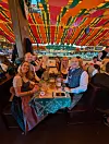 Menschen sitzen unter bunten Stoffbahnen an einer langen Tafel und halten Gläser in einem festlich dekorierten Zelt.